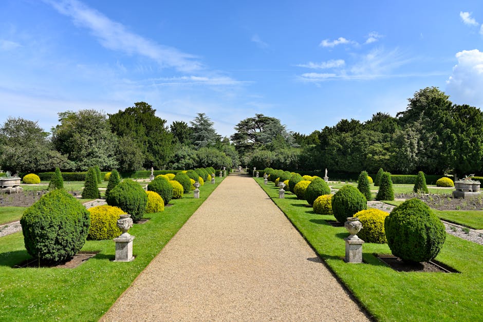 A neatly maintained garden scene showing a wooden bench positioned on a small brick-paved area alongside a lush, green lawn. The garden features a variety of plants and shrubs, including low-growing l